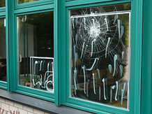 Ein Fenster weist Einschlagspuren eines Steinwurfs auf, ein benachbartes Risse, beide sind durch aufgesprhte Streifen als defekt markiert